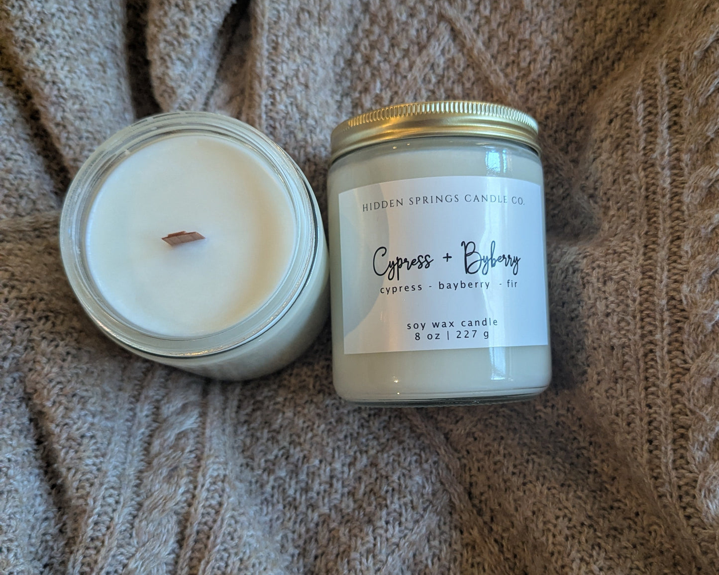 Two candles, one in a glass jar and one in a metal container, on a textured fabric background.