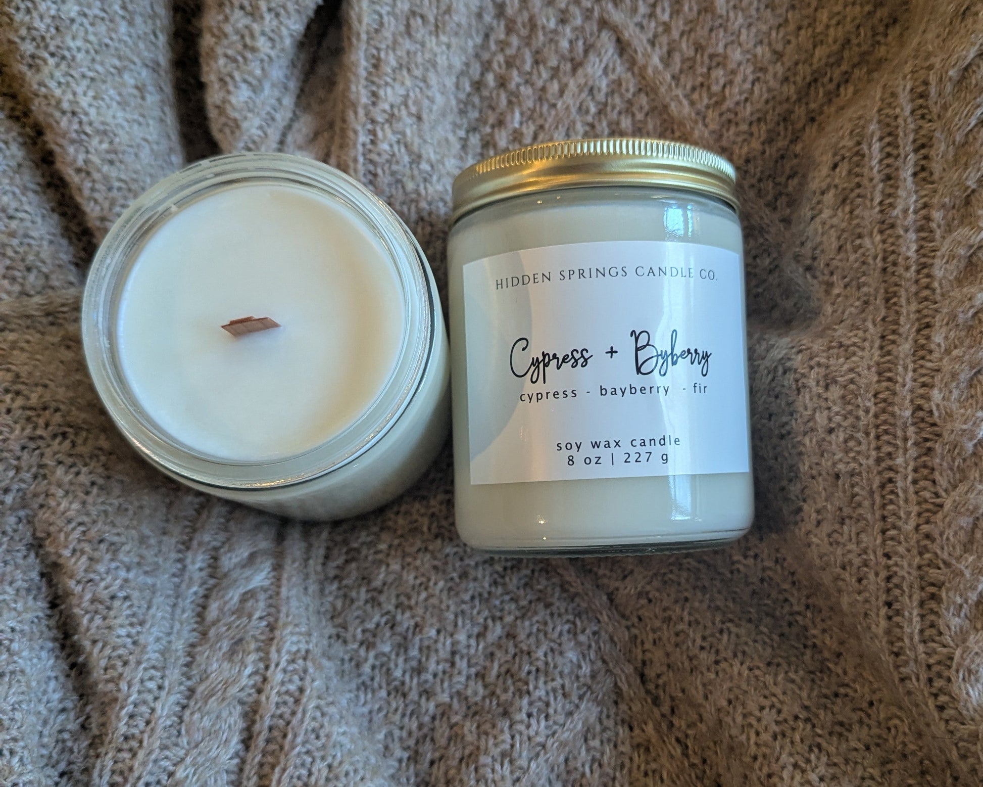 Two candles, one in a glass jar and one in a metal container, on a textured fabric background.