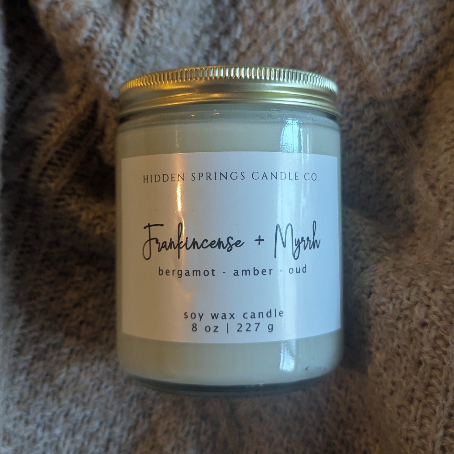 Candle jar labeled 'Frangipane + Myrrh' on a textured fabric background