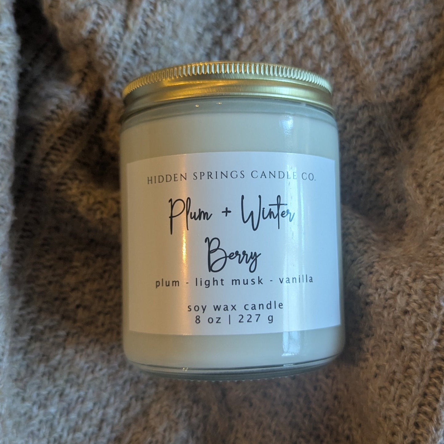 Candle jar labeled 'Plum + Water Berry' on a textured fabric background