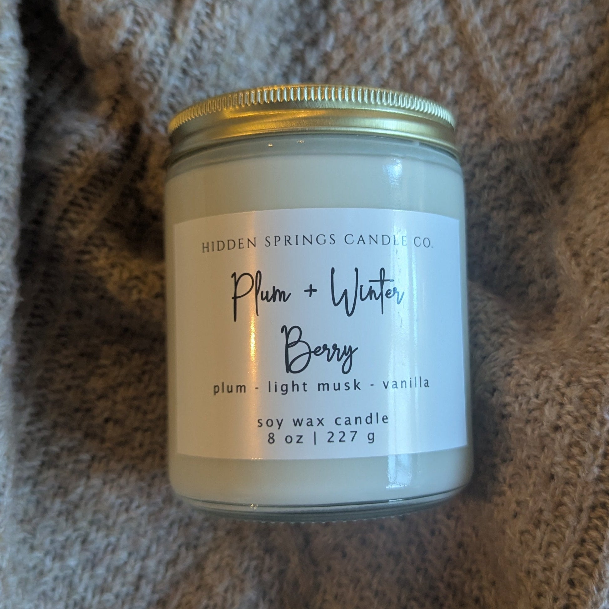 Candle jar labeled 'Plum + Water Berry' on a textured fabric background