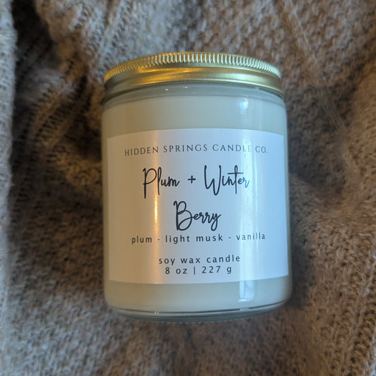 Candle jar labeled 'Plum + Water Berry' on a textured fabric background